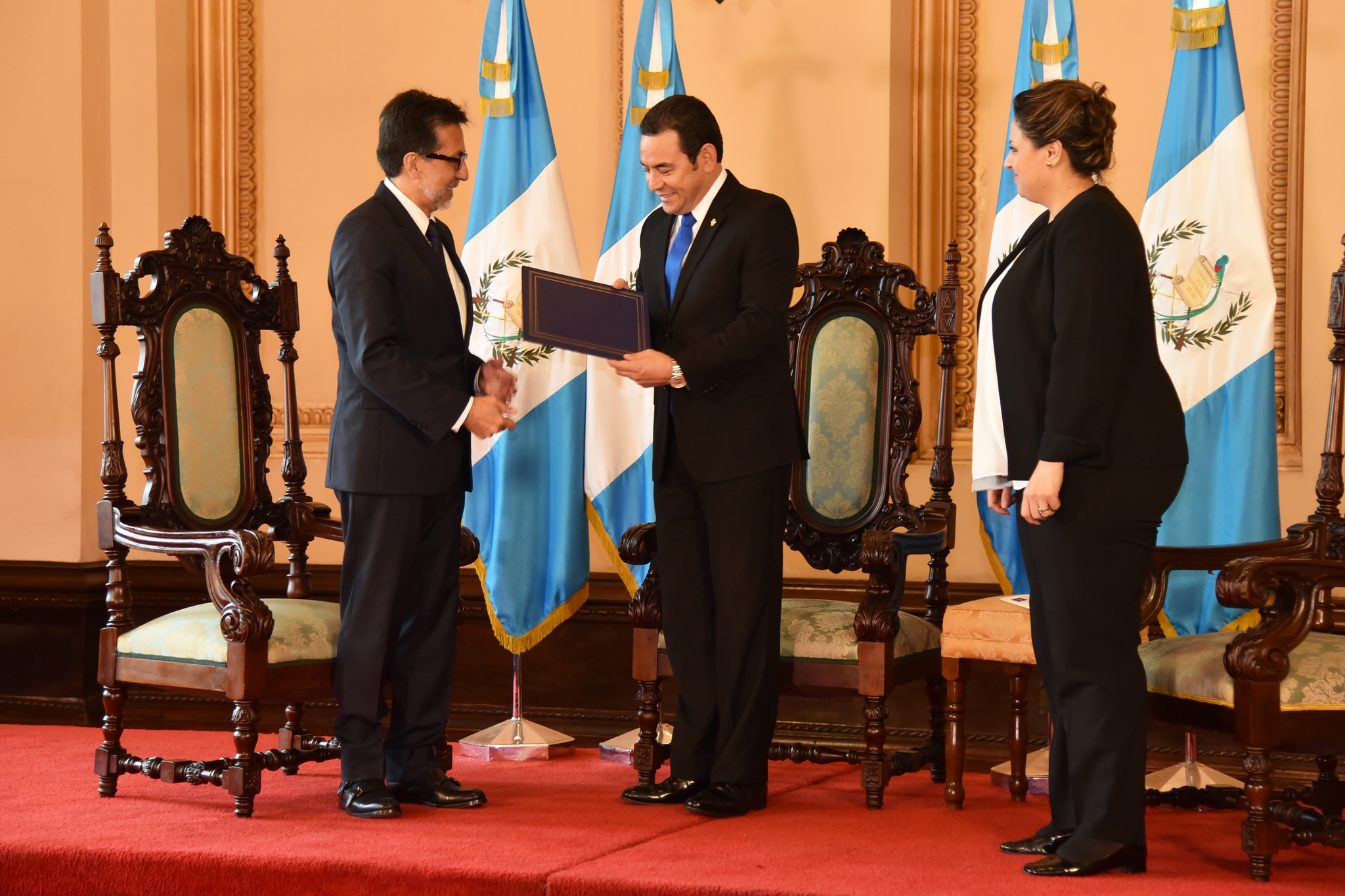 Three people stand on a dias with Guatemalan flags in the background