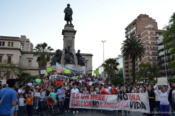 A large group of women and men gather in front of statue in a plaza.