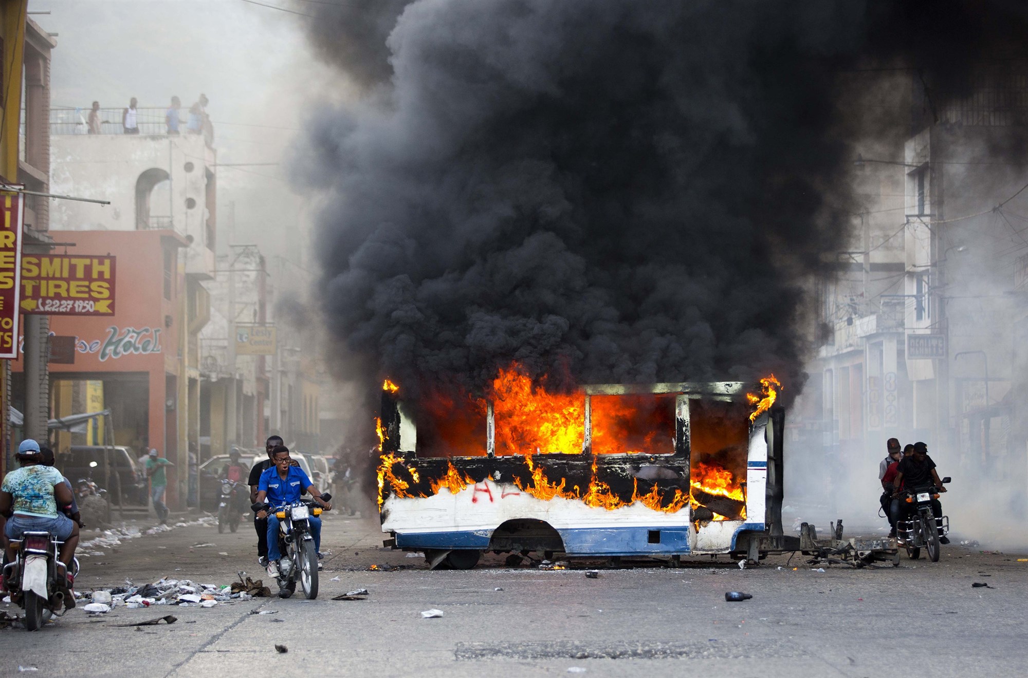 A bus burning with thick clouds of black smoke billowing above it, and two men on a motorcycle riding by.